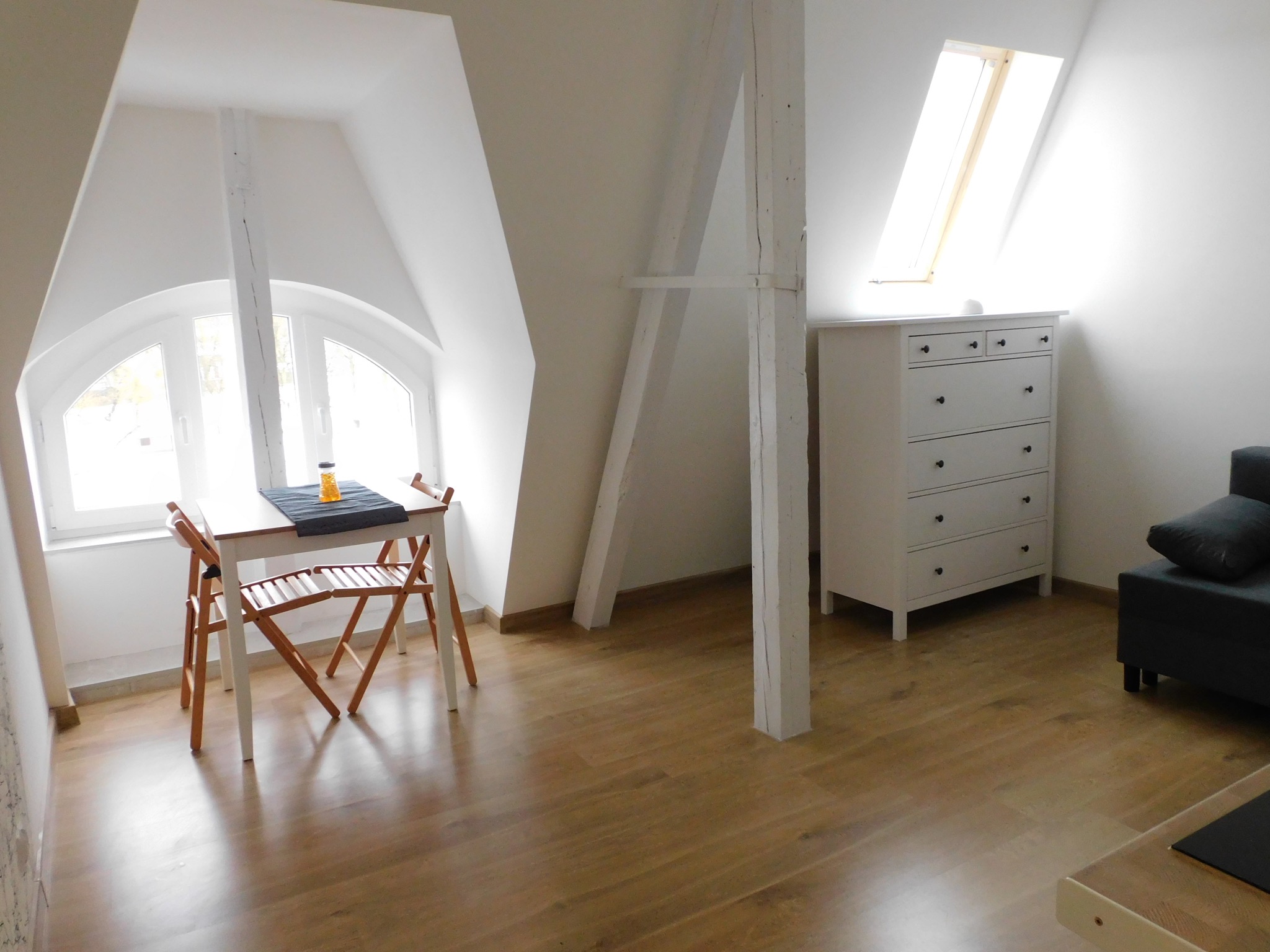 Jasne poddasze z drewnianą podłogą, białymi ścianami i stolarką; widoczny stół z krzesłami, komoda pod oknem dachowym oraz fragment sofy.