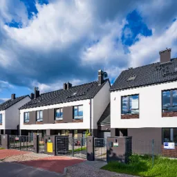 Rząd nowoczesnych domów szeregowych z białymi elewacjami, szarymi dachami i bramami wjazdowymi, na tle błękitnego nieba z dramatycznymi chmurami.