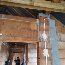 Andrzej Mida F.H.U. EL-MID,ET-MIX - Surowy stan poddasza z ceglanymi ścianami, widoczne instalacje elektryczne w białych rurkach, drewniana konstrukcja dachu i osoby w tle. Etap prac wykończeniowych.