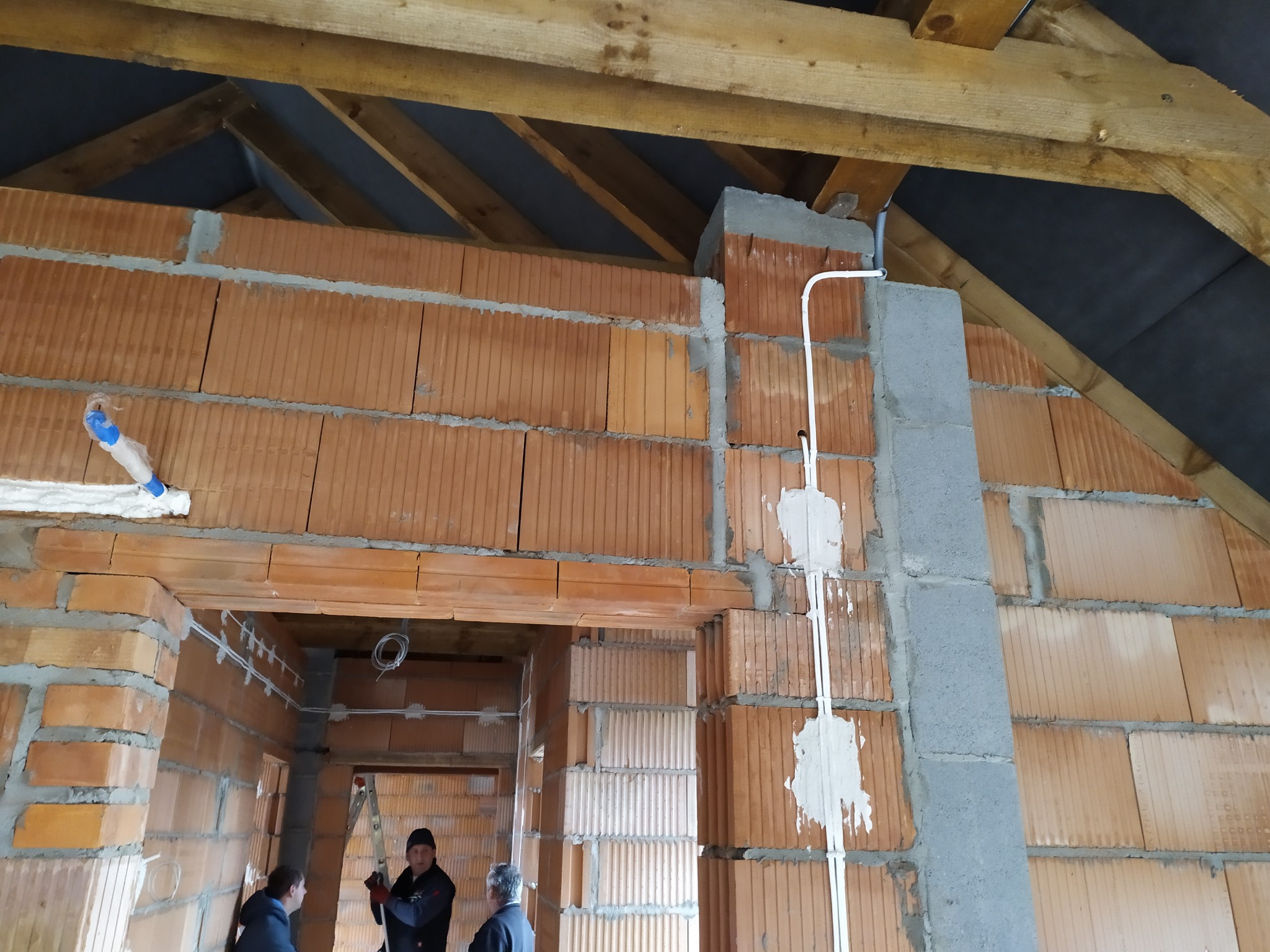 Surowy stan poddasza z ceglanymi ścianami, widoczne instalacje elektryczne w białych rurkach, drewniana konstrukcja dachu i osoby w tle. Etap prac wykończeniowych.