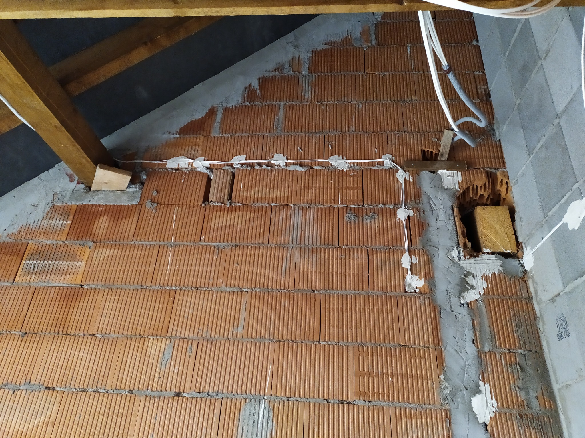 Instalacja elektryczna w trakcie montażu na ścianie z cegły dziurawki. Widoczne kable przymocowane do ściany i fragment drewnianej konstrukcji dachu.