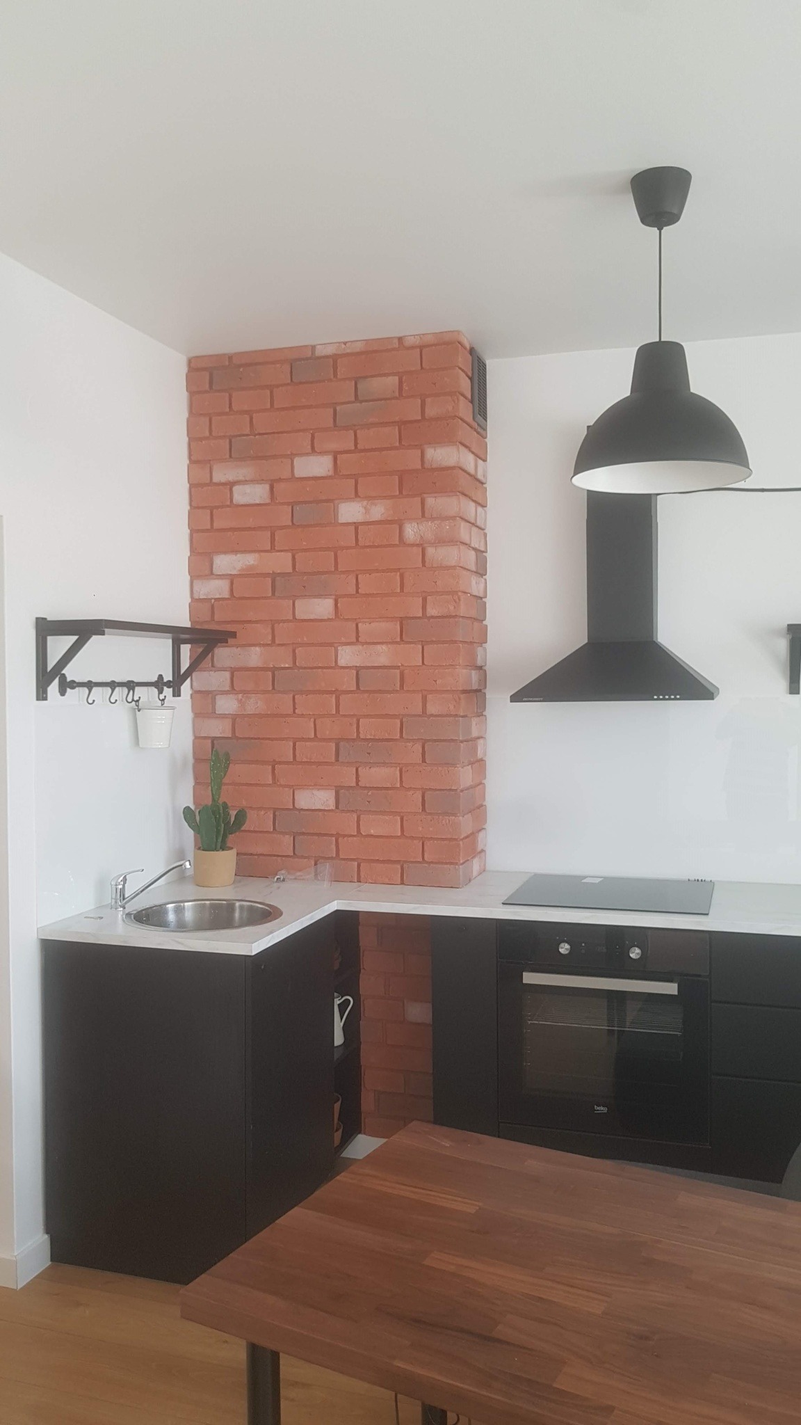 Nowoczesna kuchnia z ceglanym kominem wentylacyjnym i czarnymi szafkami w Krakowie. Minimalistyczny design, oświetlenie punktowe i drewniany stół.