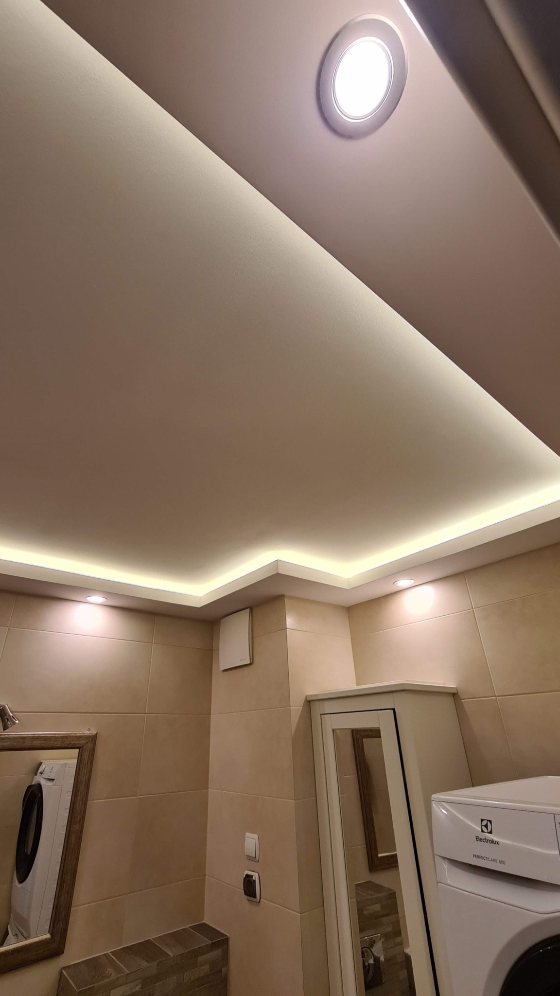 Podwieszany sufit z oświetleniem LED w łazience z beżowymi płytkami. Widoczna lampa sufitowa, listwa LED i fragment pralki Electrolux.
