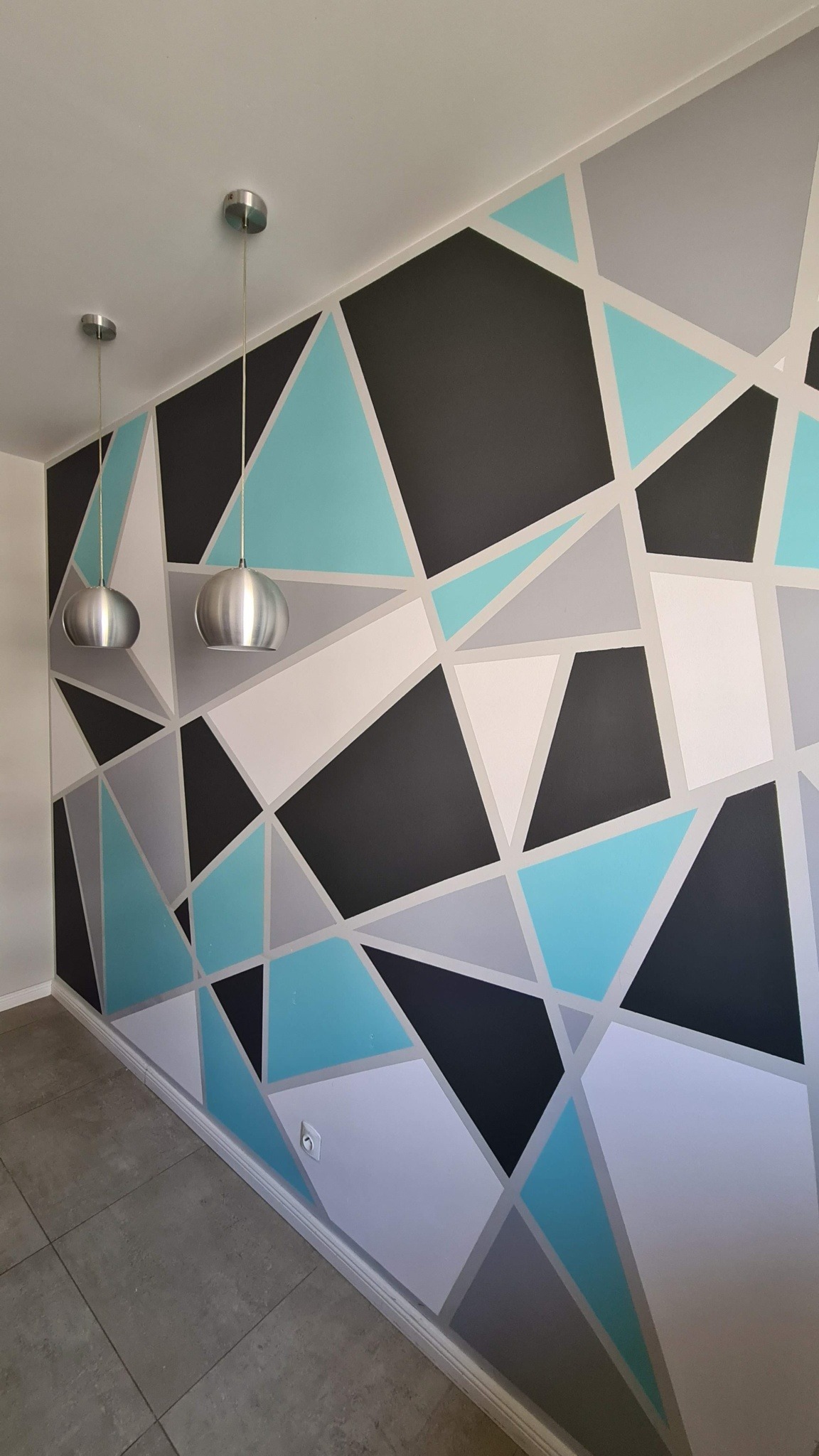 Ściana z geometrycznym wzorem w odcieniach szarości, błękitu i czerni, oświetlona przez dwie wiszące lampy z metalowymi kloszami. Widoczny fragment podłogi z szarych płytek.