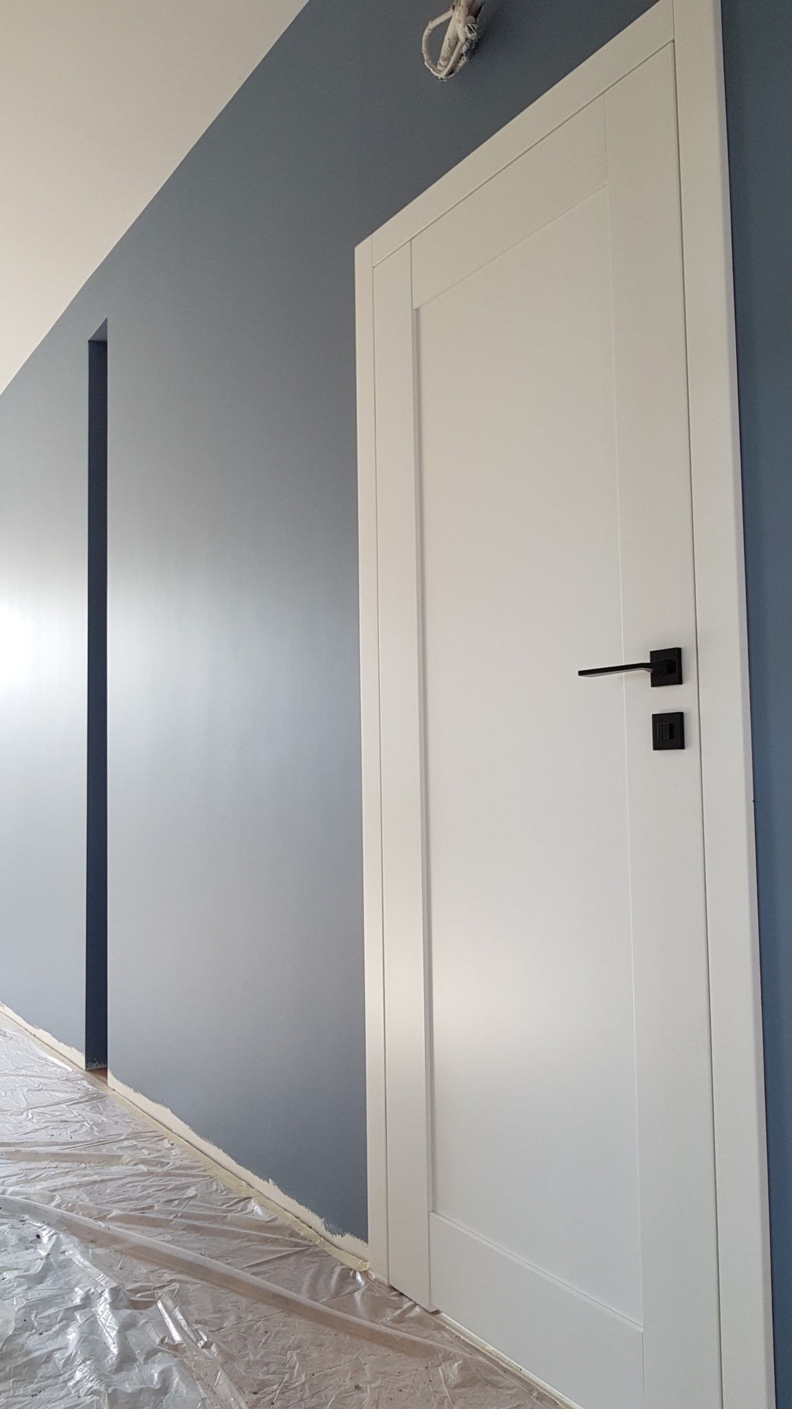 Białe drzwi z czarną klamką na tle świeżo pomalowanej, szarej ściany. Podłoga zabezpieczona folią malarską. Widoczne przewody oświetleniowe.