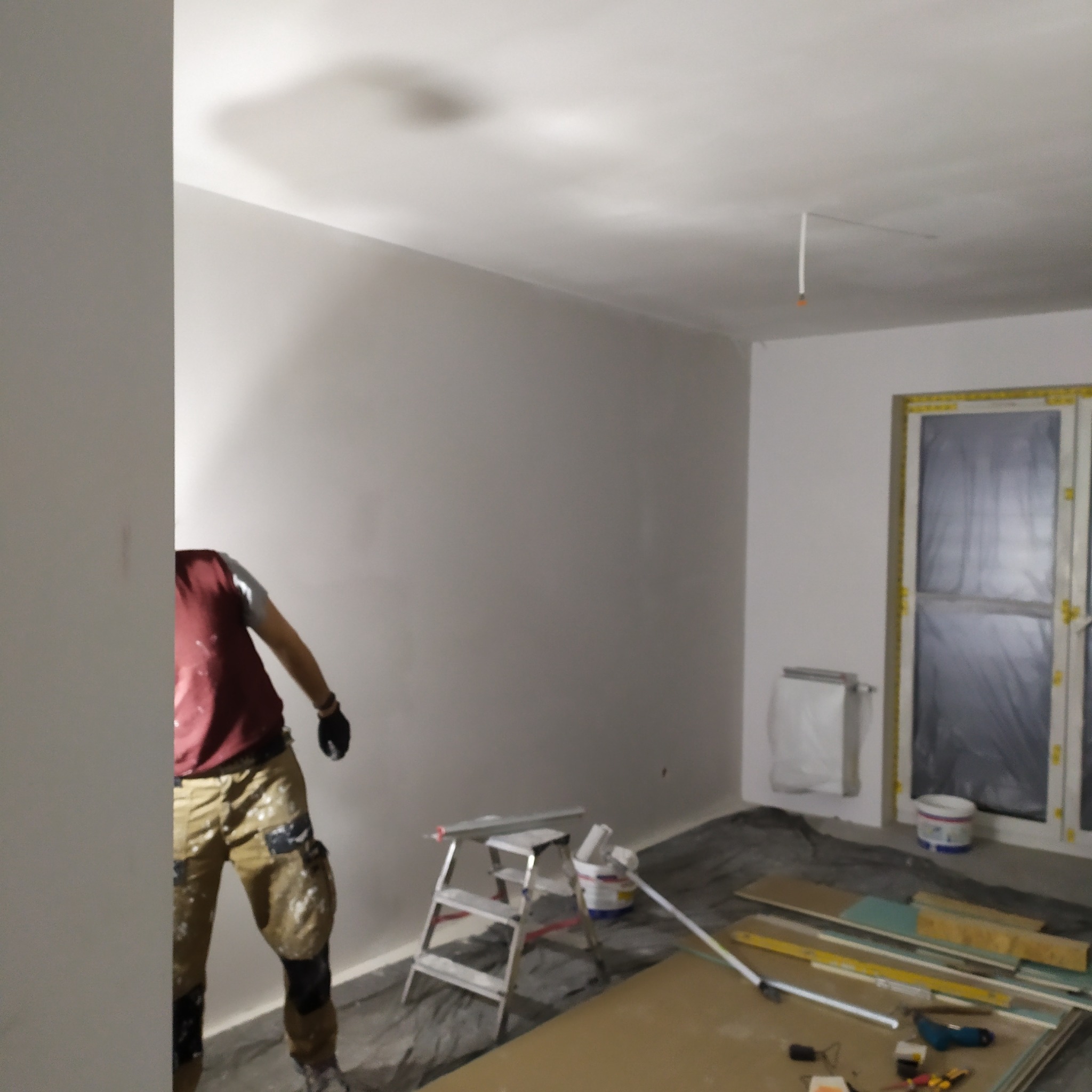 Prace wykończeniowe w pokoju: malowanie ścian na biało. Widoczny pracownik w ubraniu roboczym, drabina, narzędzia i materiały budowlane. Okno zabezpieczone folią.