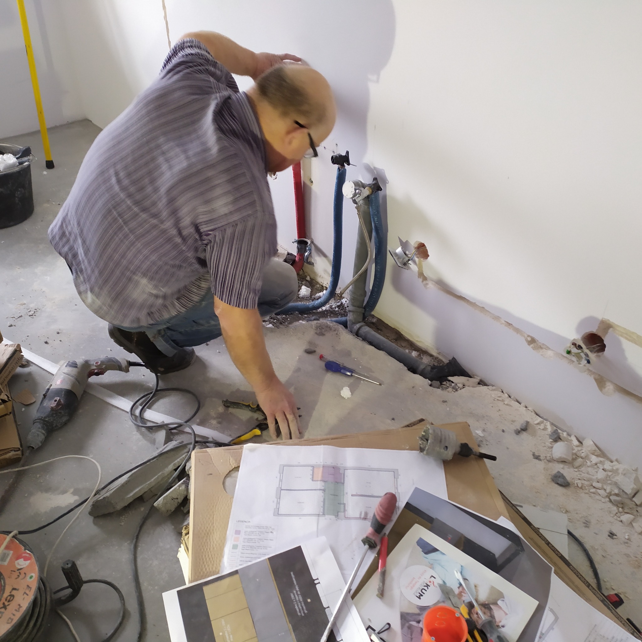 Instalator w trakcie pracy nad instalacją wodną w pomieszczeniu w stanie surowym. Widoczne narzędzia, plany i elementy instalacji.