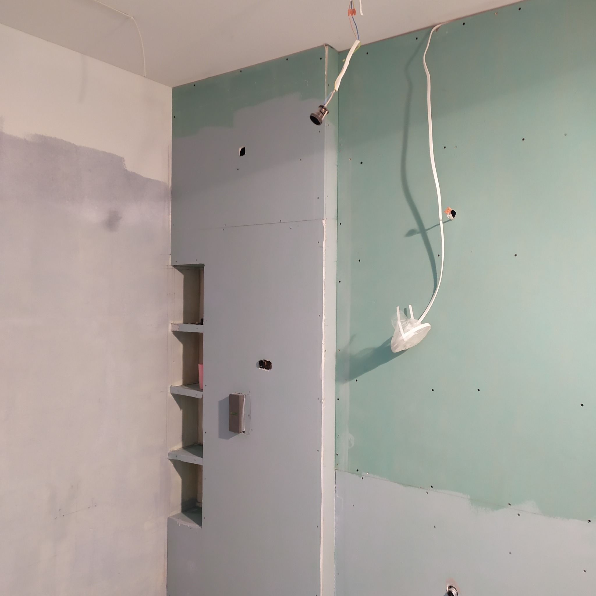 Ściana z płyt gipsowo-kartonowych w trakcie wykończenia, widoczne kable elektryczne i wnęka z półkami. Etap prac wykończeniowych w pomieszczeniu.