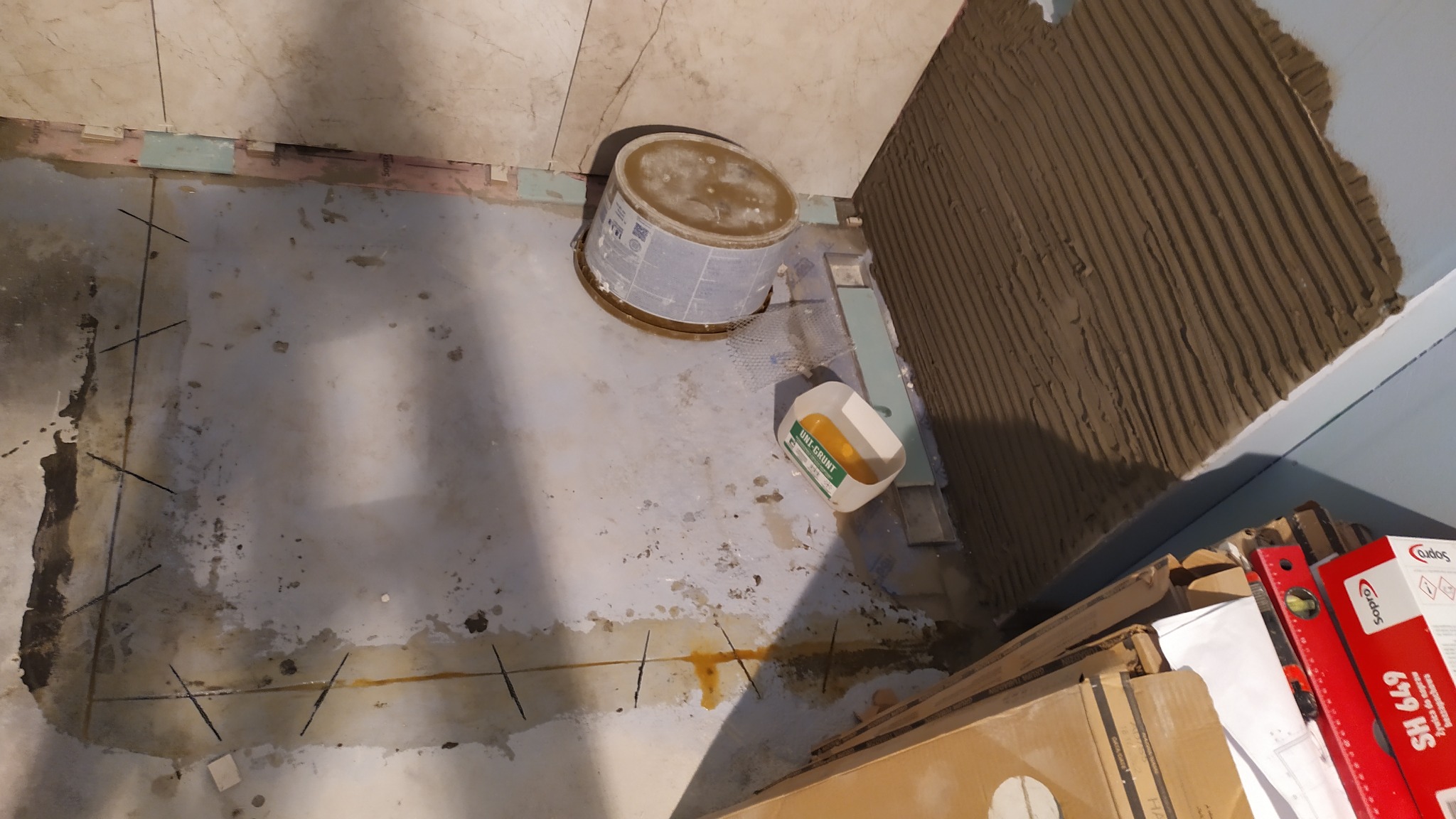 Przygotowanie podłoża pod płytki w łazience: gruntowanie, wyznaczanie linii cięcia. Widoczne narzędzia i materiały budowlane. Prace remontowe w toku.