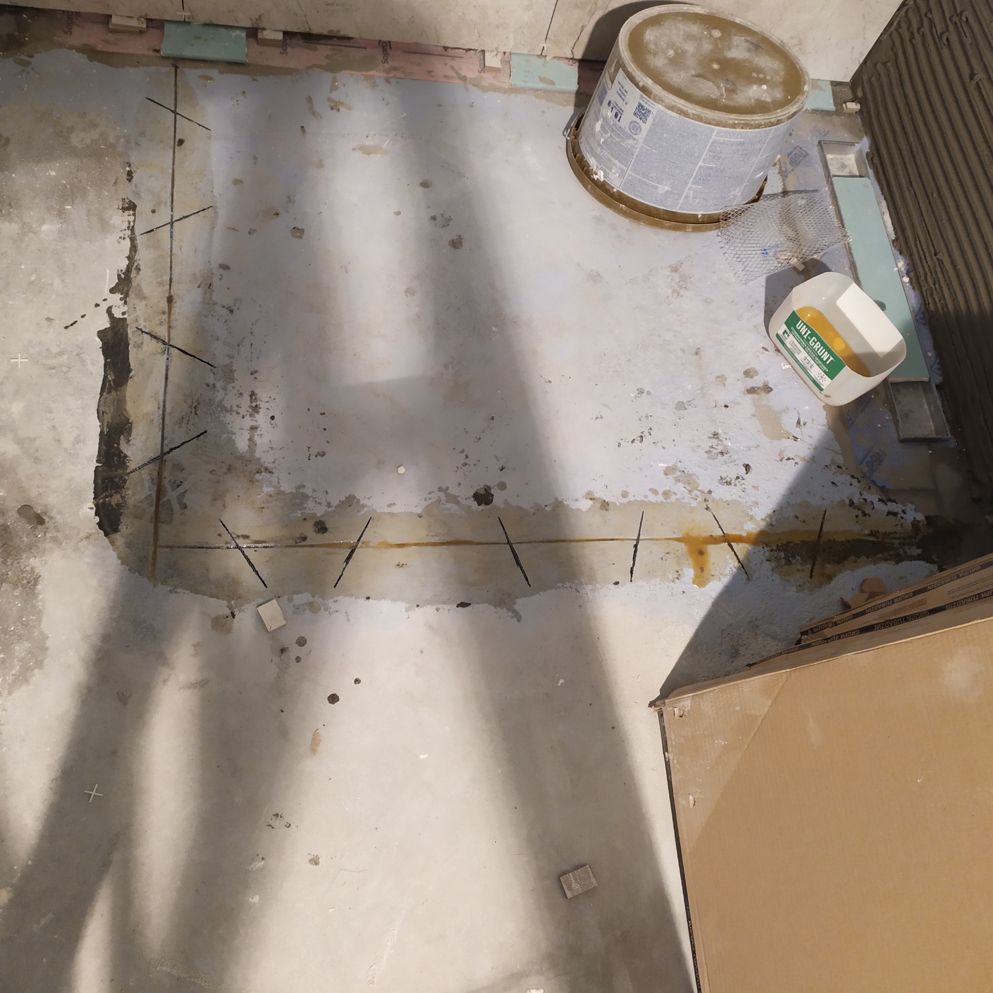 Przygotowanie podłoża pod płytki: gruntowanie i wyznaczanie linii cięcia. Widoczne narzędzia i materiały budowlane. Ujęcie z góry, surowy stan przed wykończeniem.