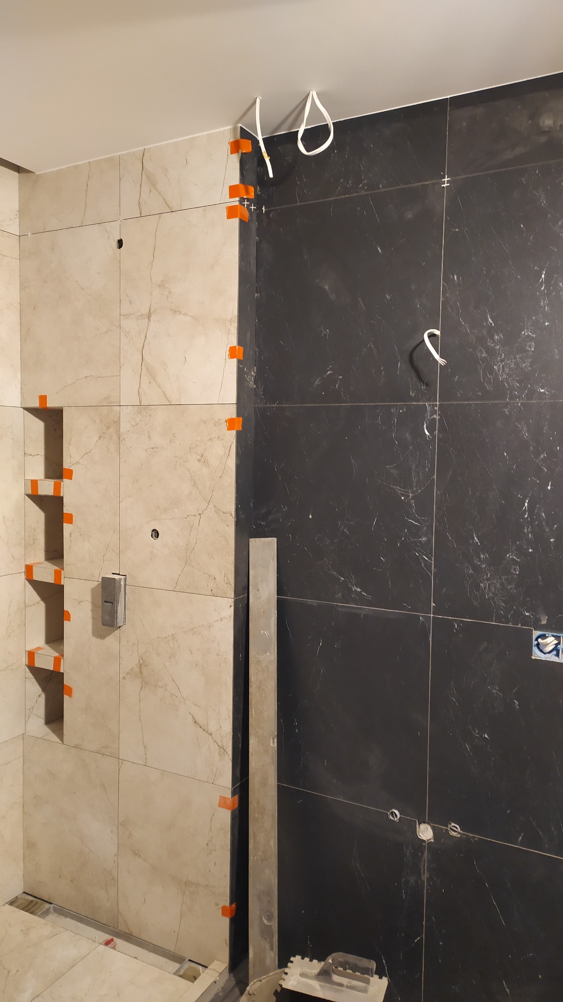 Układanie płytek w łazience w Krakowie. Widoczne płytki w odcieniach beżu i czerni, z pomarańczowymi krzyżykami dystansowymi. Częściowo wykończona ściana z wnęką.