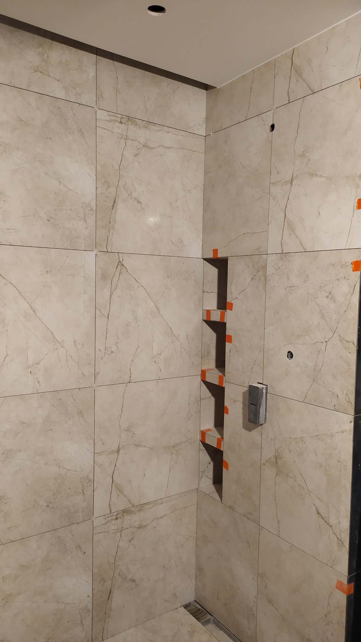 Beżowe płytki imitujące marmur w kabinie prysznicowej z wnęką na półki, zabezpieczone pomarańczową taśmą. Widoczny odpływ liniowy i gniazdko elektryczne.