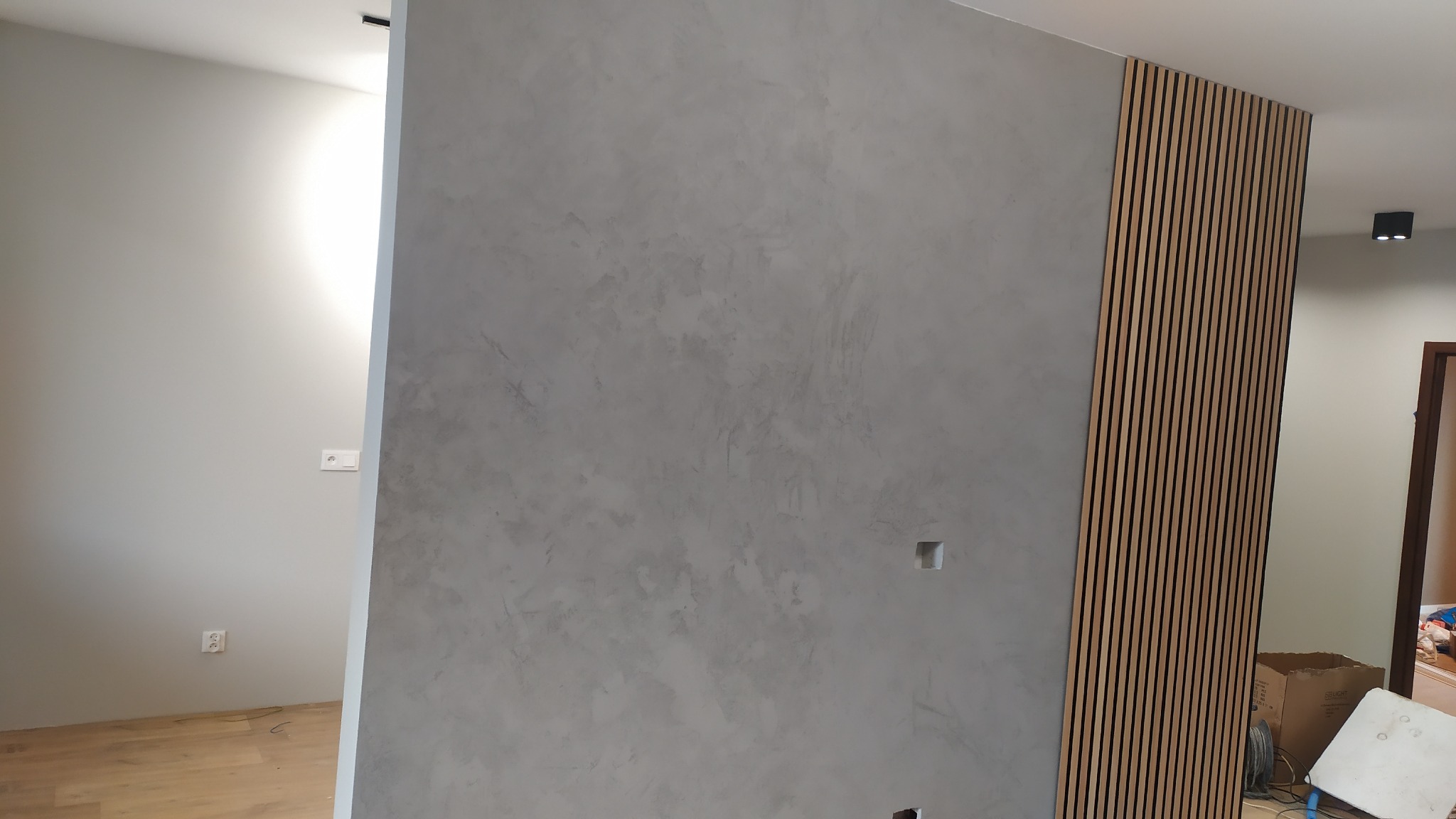 Ściana z szarą, dekoracyjną fakturą i drewnianymi lamelami. Widoczne gniazdka elektryczne. Nowoczesne wykończenie wnętrza z akcentem naturalnego drewna.