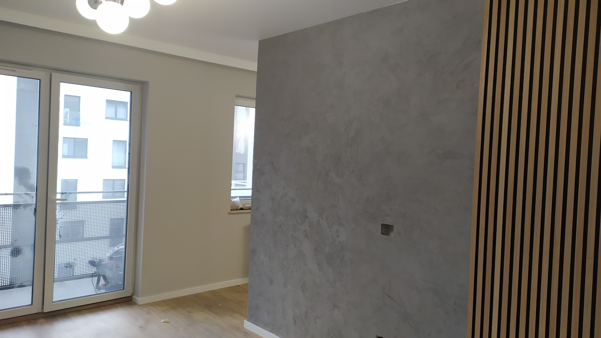 Szara ściana z efektem dekoracyjnym i drewniane lamele w jasnym wnętrzu z oknem balkonowym. Widoczny fragment podłogi i sufitu z oświetleniem.