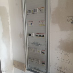 IMS INSTALACJE I BUDOWA - Szara skrzynka rozdzielcza z bezpiecznikami zamontowana na ścianie w trakcie remontu, widoczne notatki ołówkiem na ścianie i surowe wykończenie.