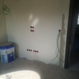 IMS INSTALACJE I BUDOWA - Wnętrze w trakcie prac wykończeniowych: widoczne puszki elektryczne w ścianie, wystające przewody, torba z materiałem izolacyjnym i parkiet ułożony w jodełkę.