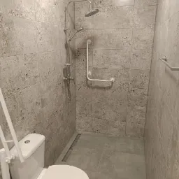 Wykończona łazienka z szarymi, wzorzystymi płytkami, kabiną prysznicową typu walk-in z odpływem liniowym, chromowaną baterią i deszczownicą oraz uchwytem dla niepełnosprawnych; toaleta z dodatkowym...