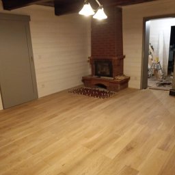 IMS INSTALACJE I BUDOWA - Jasna drewniana podłoga w pokoju z kominkiem z cegły w Kędzierzynie-Koźlu, widoczne przygotowane płytki podłogowe przed kominkiem, ściany wykończone boazerią, w tle widoczne schody i drabina.