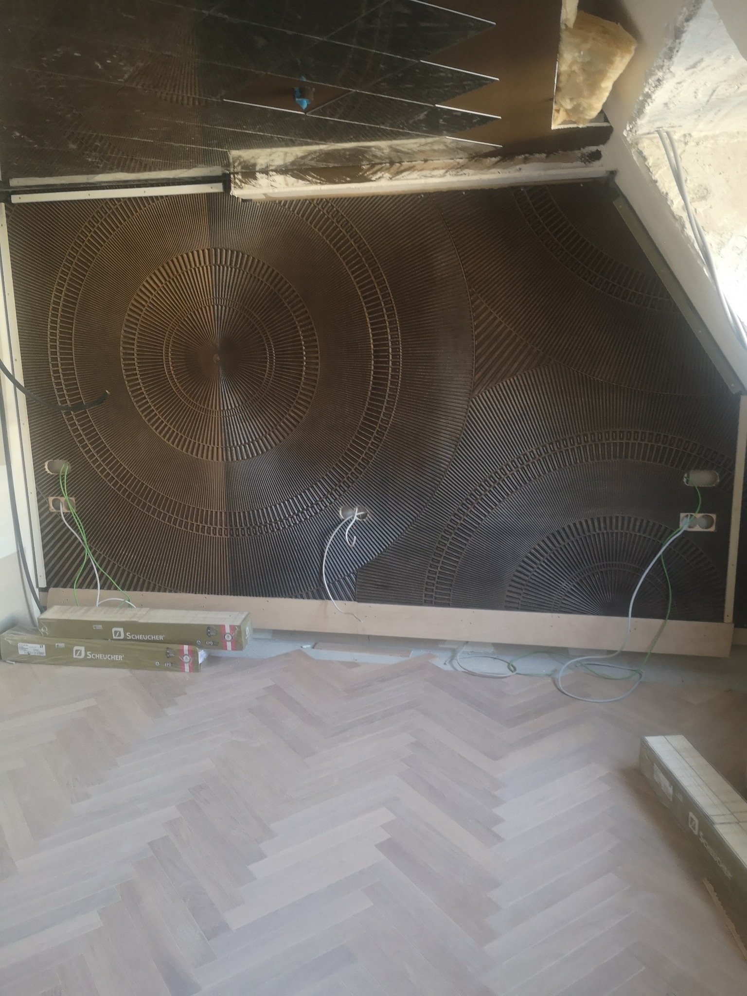 Ściana z dekoracyjnymi panelami z motywem koncentrycznych okręgów, widoczne gniazdka elektryczne z wystającymi przewodami, podłoga ułożona w jodełkę, fragment sufitu podwieszanego w trakcie montażu.