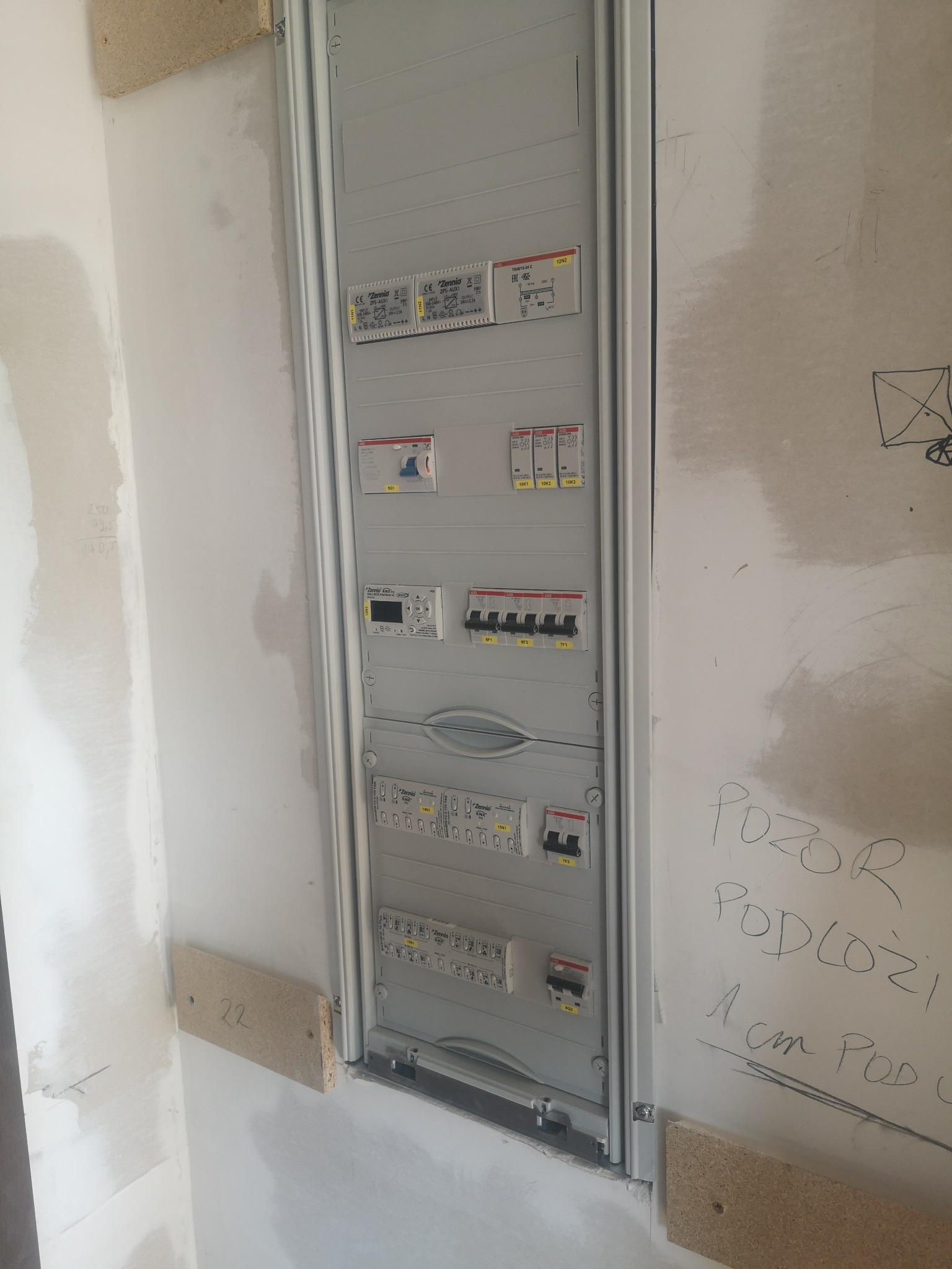 Szara skrzynka rozdzielcza z bezpiecznikami zamontowana na ścianie w trakcie remontu, widoczne notatki ołówkiem na ścianie i surowe wykończenie.
