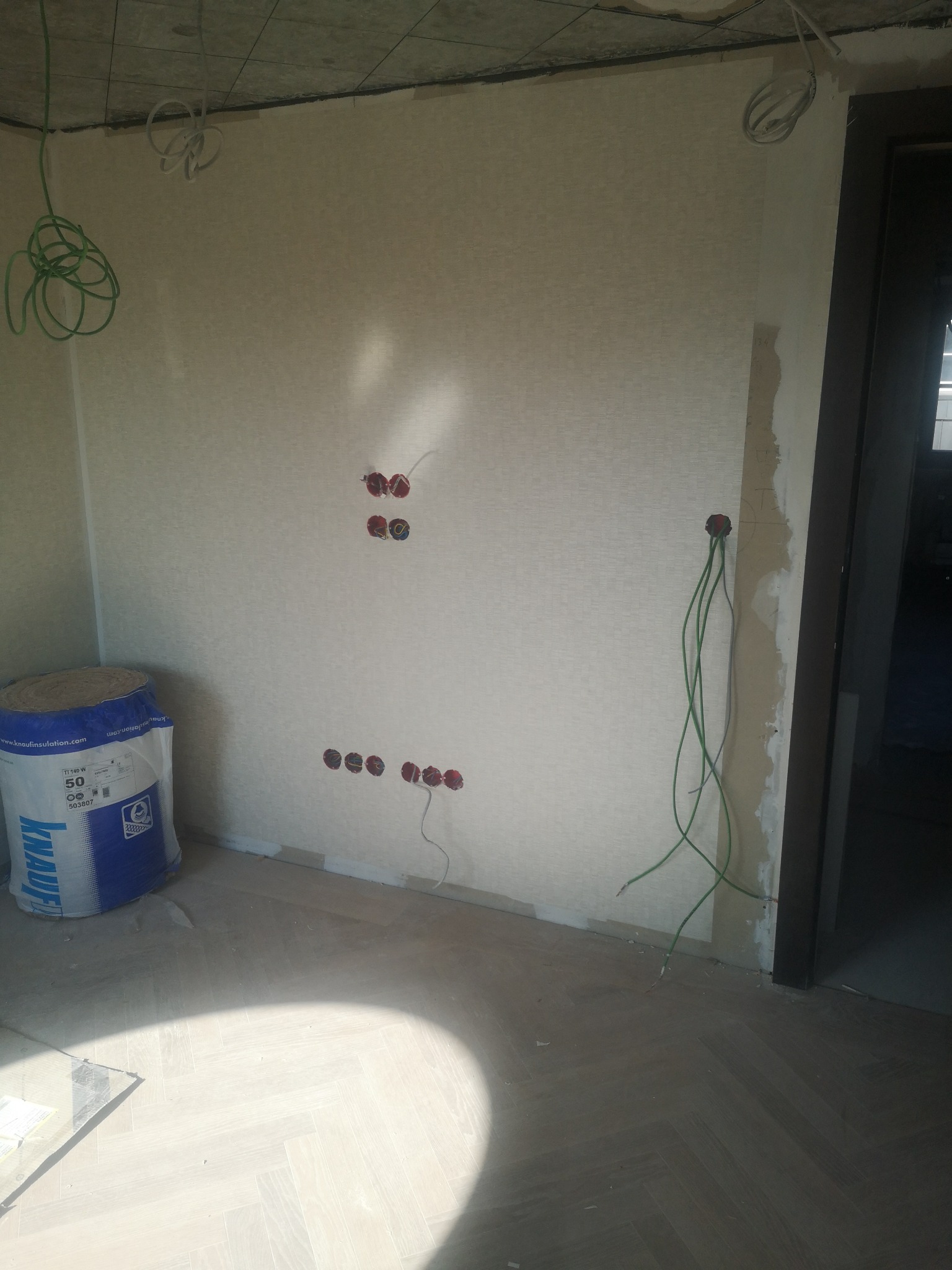 Wnętrze w trakcie prac wykończeniowych: widoczne puszki elektryczne w ścianie, wystające przewody, torba z materiałem izolacyjnym i parkiet ułożony w jodełkę.