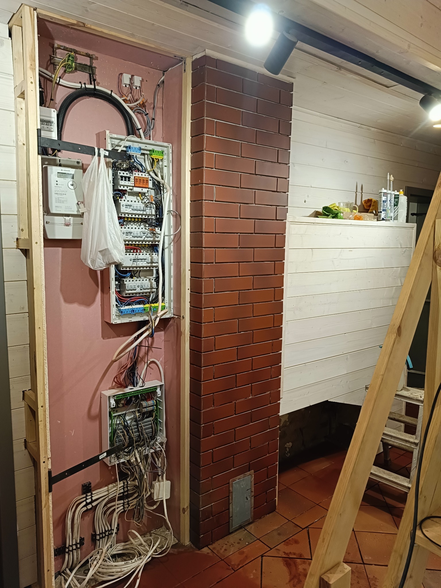 Rozdzielnica elektryczna w trakcie montażu w drewnianej ramie, widoczne liczniki, kable i zabezpieczenia, ściana pomalowana na różowo, obok mur z cegły, w tle drewniana boazeria i drabina.