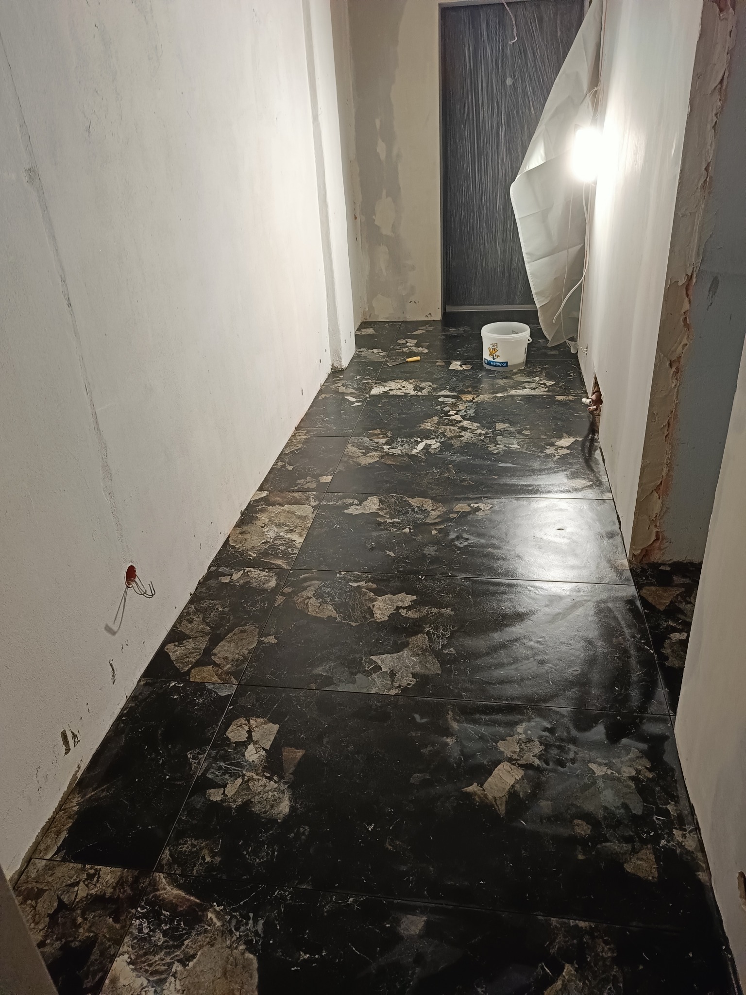 Korytarz w trakcie remontu: czarne płytki podłogowe z marmurkowym wzorem, ściany w trakcie malowania, zabezpieczone folią drzwi, wiadro farby i wystające kable elektryczne ze ściany.