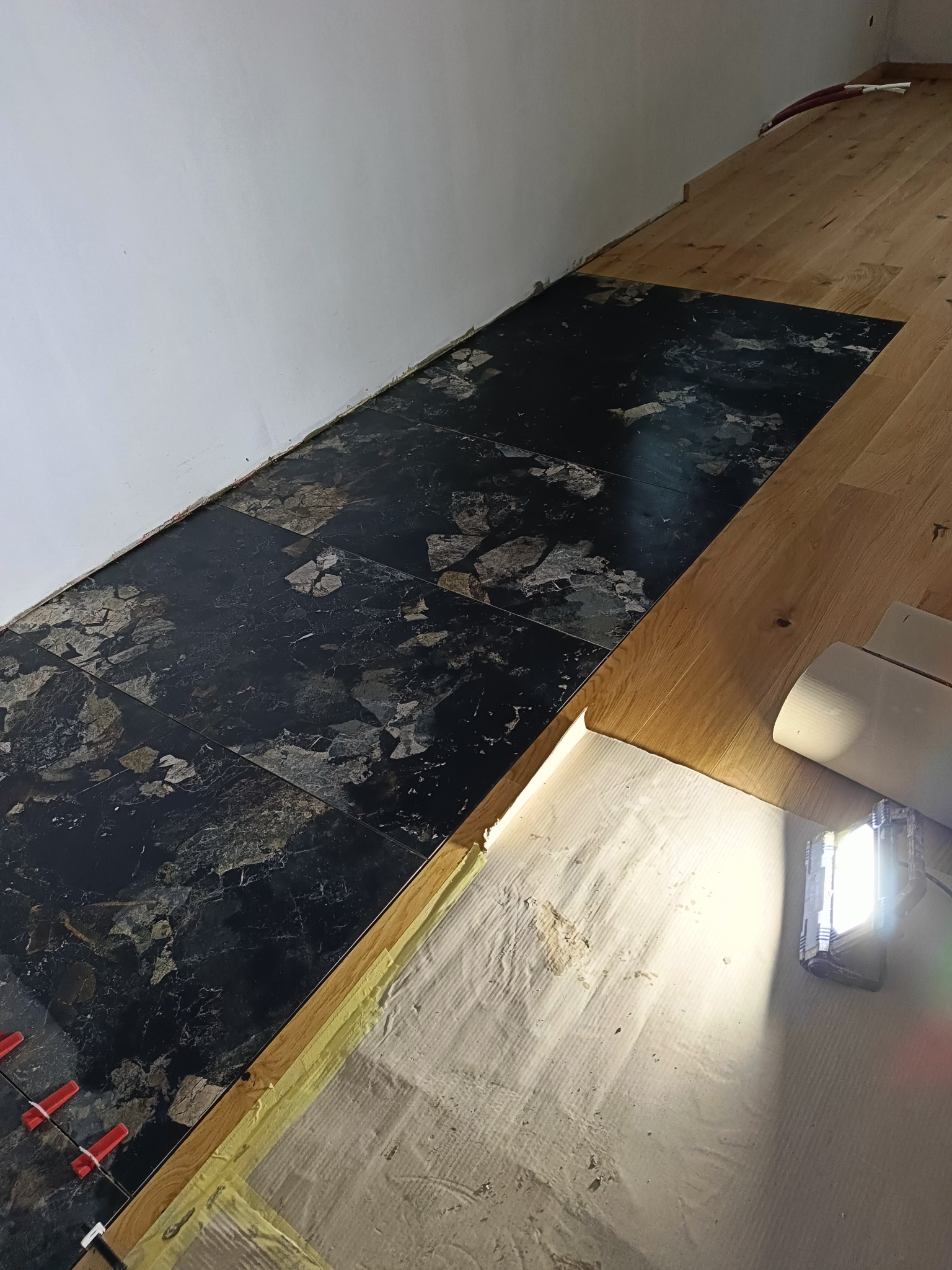 Fragment podłogi z ułożonymi czarnymi płytkami imitującymi marmur, z widocznymi czerwonymi klinami do poziomowania i żółtą taśmą zabezpieczającą, sąsiadujący z fragmentem drewnianej podłogi...