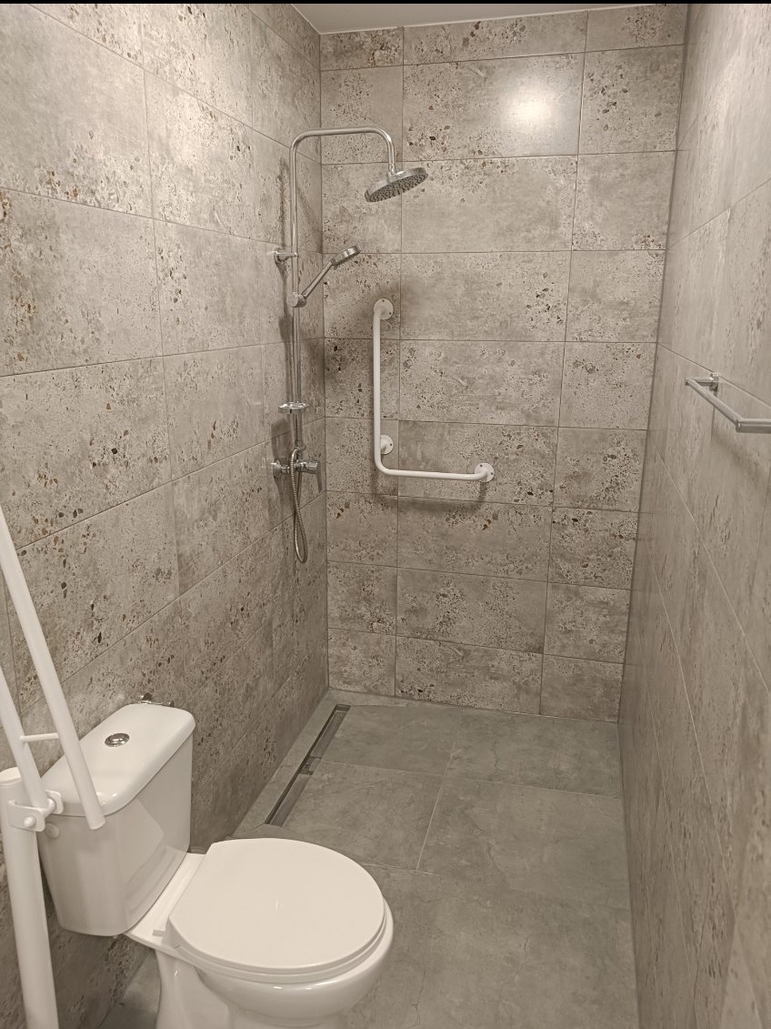 Wykończona łazienka z szarymi, wzorzystymi płytkami, kabiną prysznicową typu walk-in z odpływem liniowym, chromowaną baterią i deszczownicą oraz uchwytem dla niepełnosprawnych; toaleta z dodatkowym...