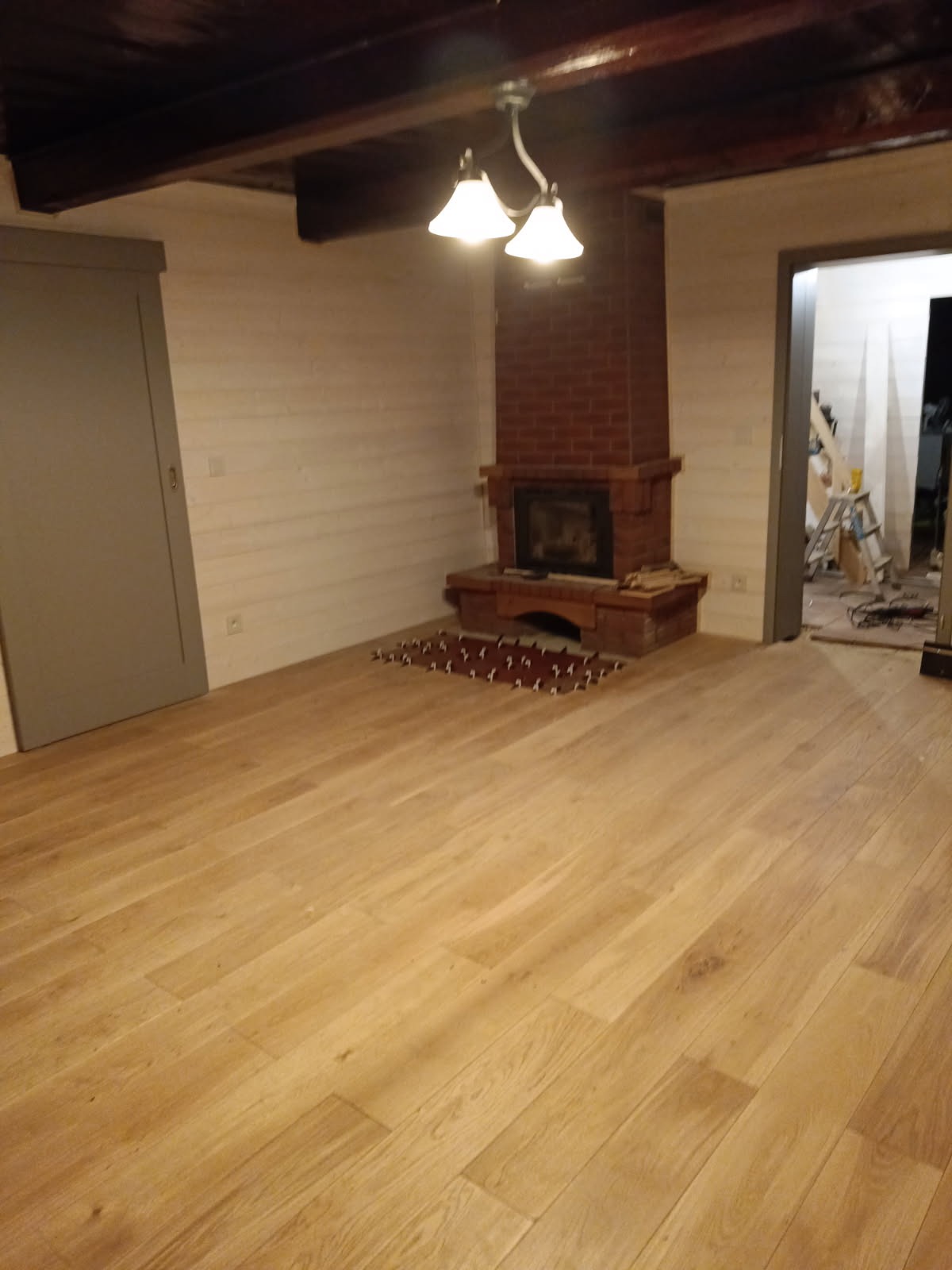Jasna drewniana podłoga w pokoju z kominkiem z cegły w Kędzierzynie-Koźlu, widoczne przygotowane płytki podłogowe przed kominkiem, ściany wykończone boazerią, w tle widoczne schody i drabina.