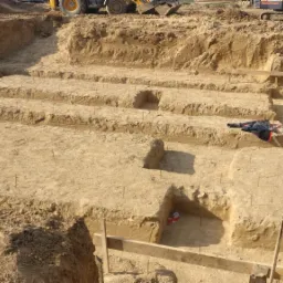 Wykopane rowy fundamentowe z wystającymi prętami zbrojeniowymi, w tle sprzęt budowlany, na ziemi leży kurtka robocza.