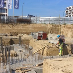 MALMAC Sp. z o.o - Prace ziemne pod fundamenty budynku wielorodzinnego, widoczne zbrojenia i szalunki, pracownik w kasku i odzieży roboczej przy wykopie, w tle budynki mieszkalne.