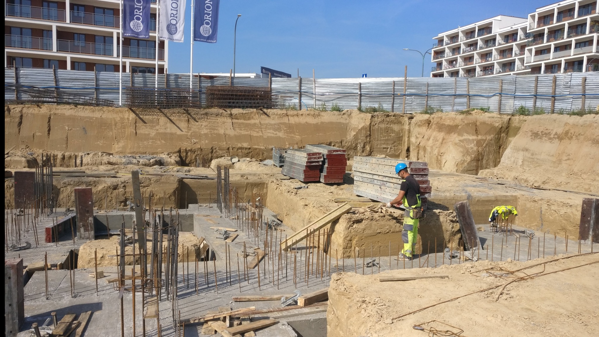 Prace ziemne pod fundamenty budynku wielorodzinnego, widoczne zbrojenia i szalunki, pracownik w kasku i odzieży roboczej przy wykopie, w tle budynki mieszkalne.