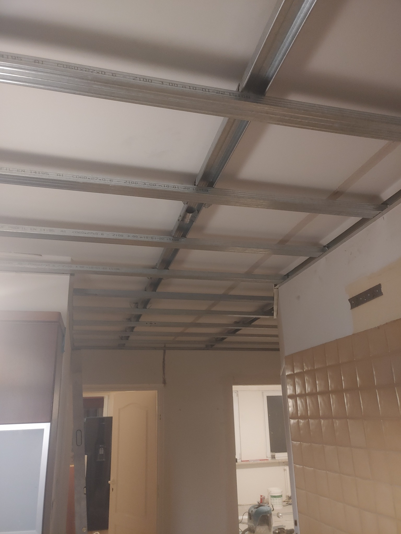 Montaż metalowej konstrukcji sufitu podwieszanego z widocznymi profilami CD60 i płytami gipsowo-kartonowymi, widok z dołu, w tle fragment pomieszczenia z beżowymi płytkami na ścianie i widocznym...