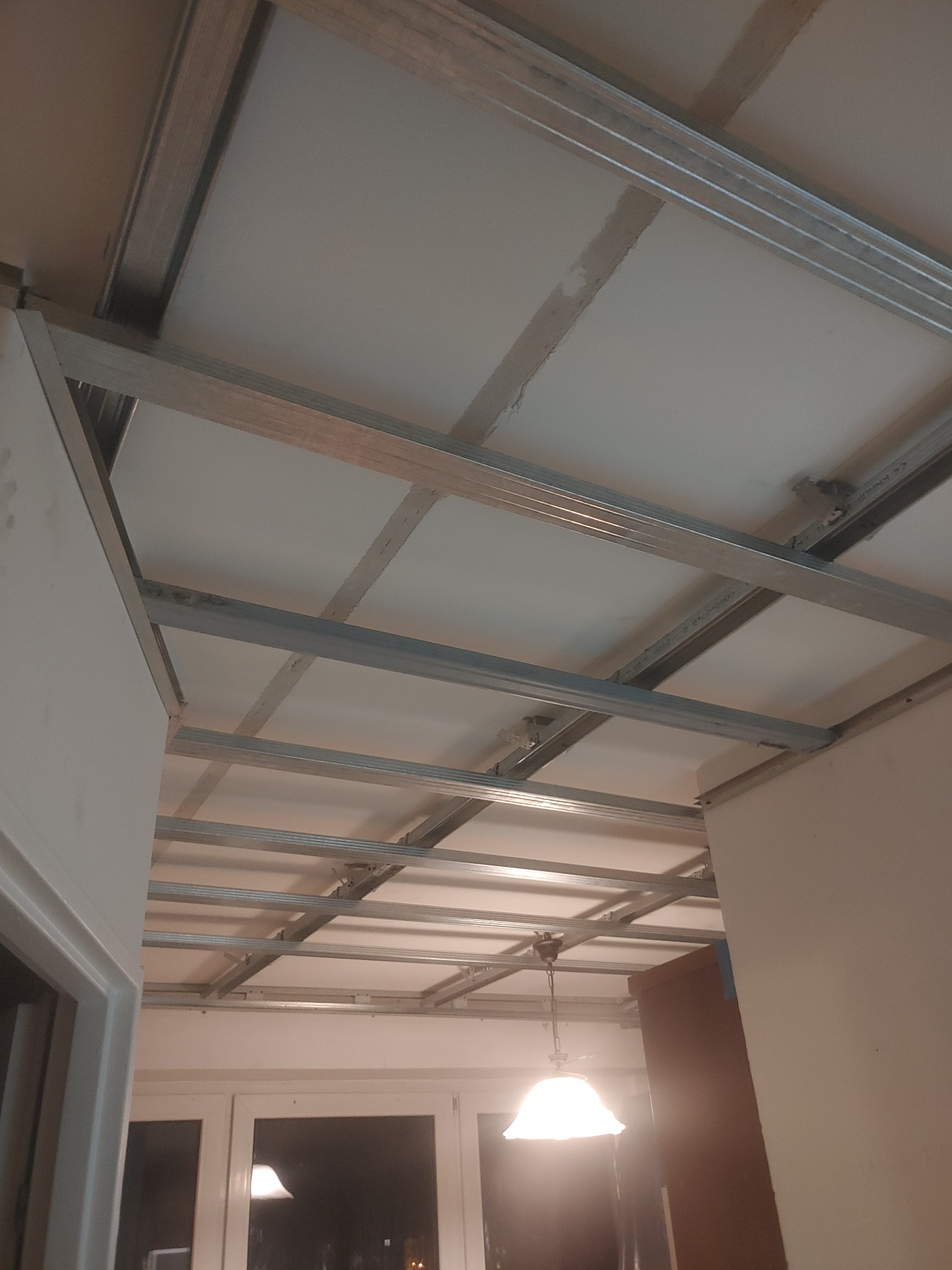 Stelaż sufitu podwieszanego z płyt gipsowo-kartonowych, widok z dołu, widoczne profile metalowe i płyty, w tle okno i lampa.