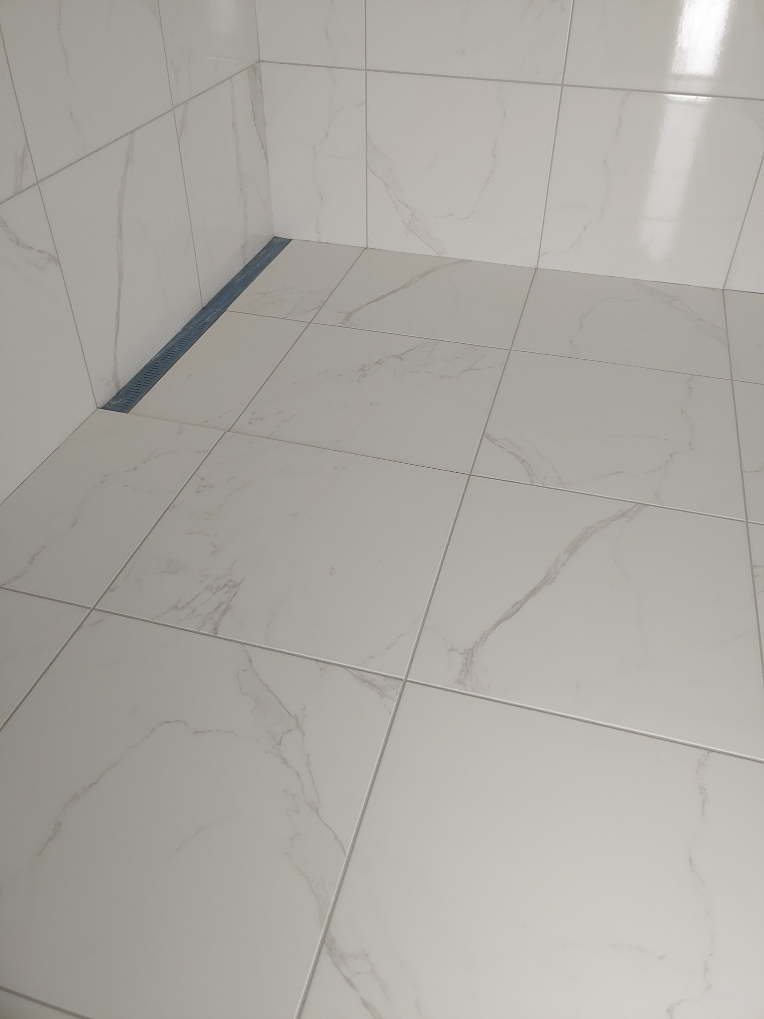 Nowoczesny odpływ liniowy w narożniku kabiny prysznicowej wyłożonej białymi płytkami imitującymi marmur z subtelnym szarym żyłkowaniem.