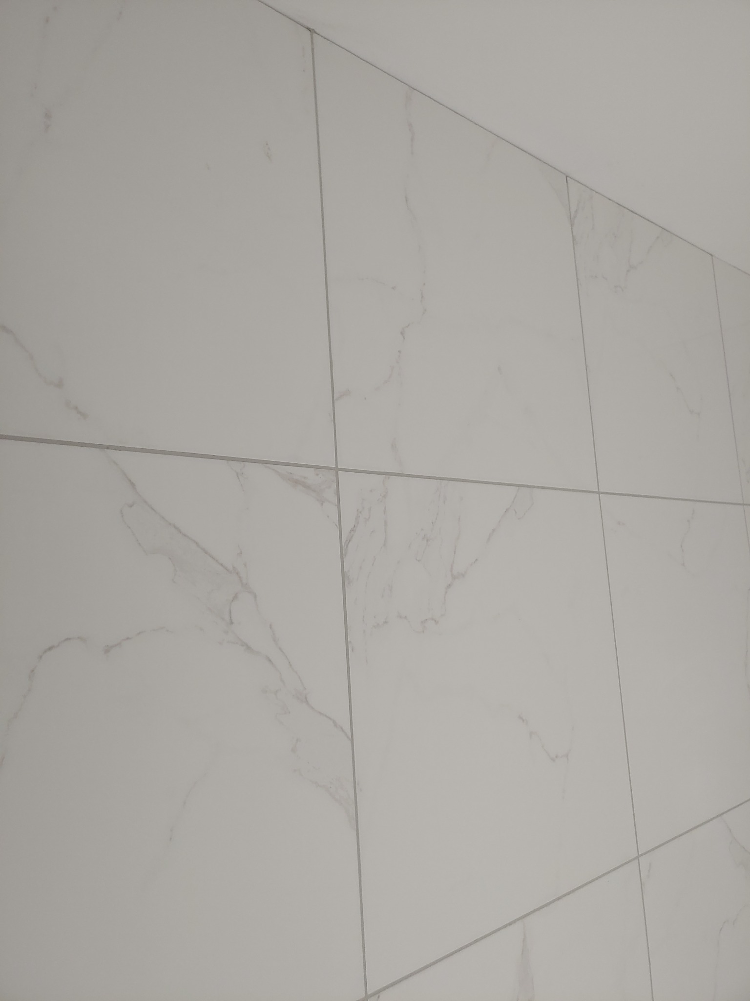 Ściana wyłożona białymi płytkami imitującymi marmur, z widocznymi szarymi fugami, sfotografowana pod kątem.