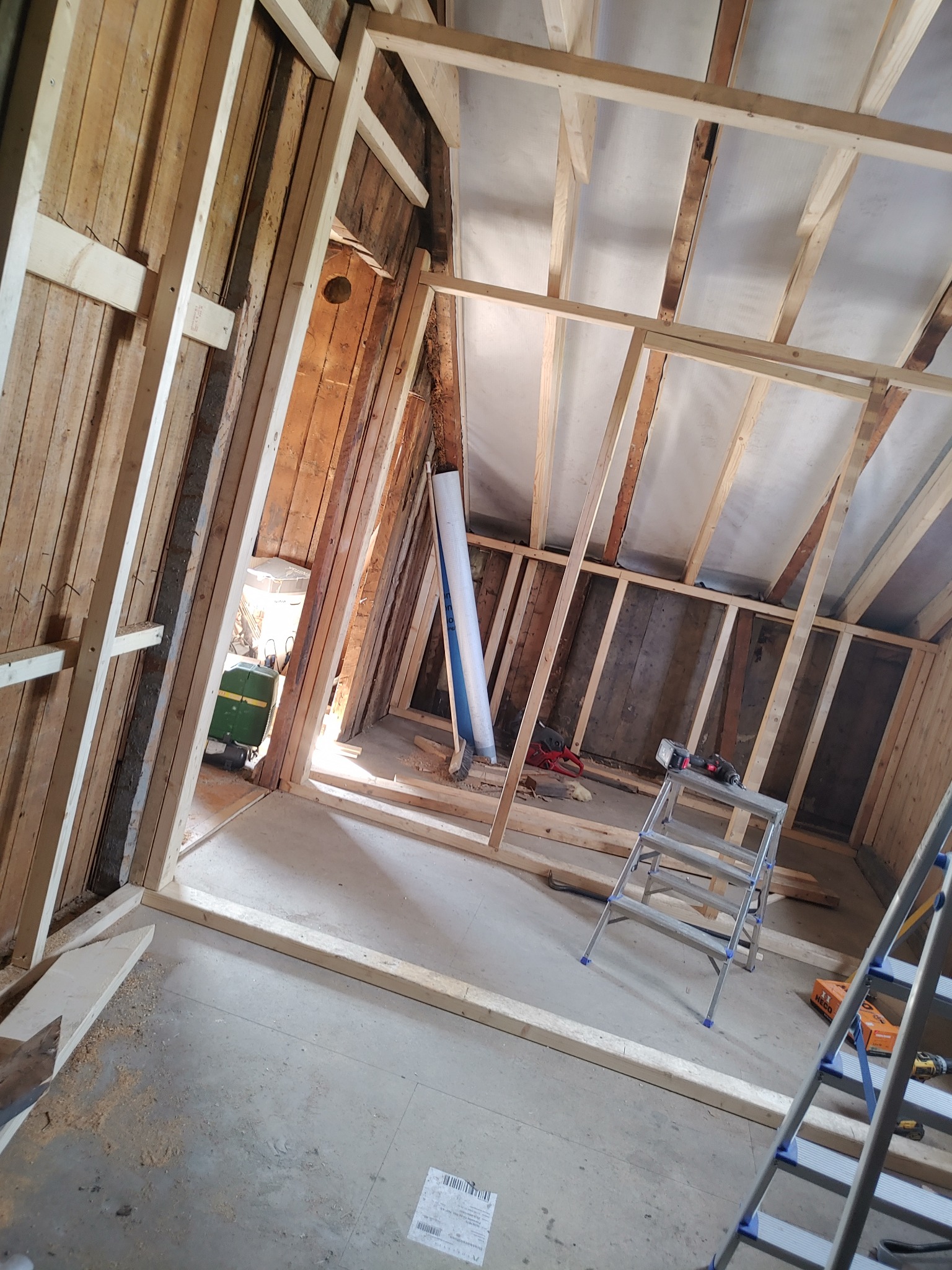Konstrukcja szkieletowa poddasza z drewnianych belek, widoczne narzędzia i drabina, częściowo ocieplony dach folią, surowe drewno na ścianach.