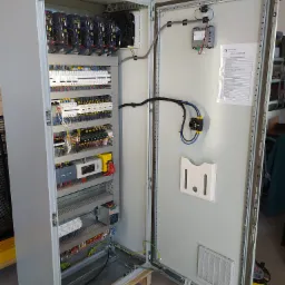 Otwarta szafa sterownicza z widocznymi modułami sterującymi, wentylatorami, okablowaniem i listwą zasilającą, z dokumentacją techniczną na wewnętrznej stronie drzwi.