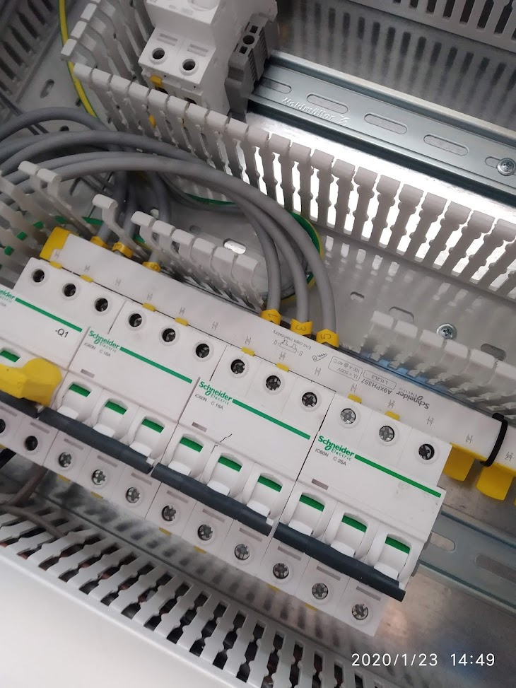 Rozdzielnica elektryczna z zamontowanymi bezpiecznikami Schneider Electric, widoczne szare przewody z żółtymi oznaczeniami podłączone do aparatury modułowej.