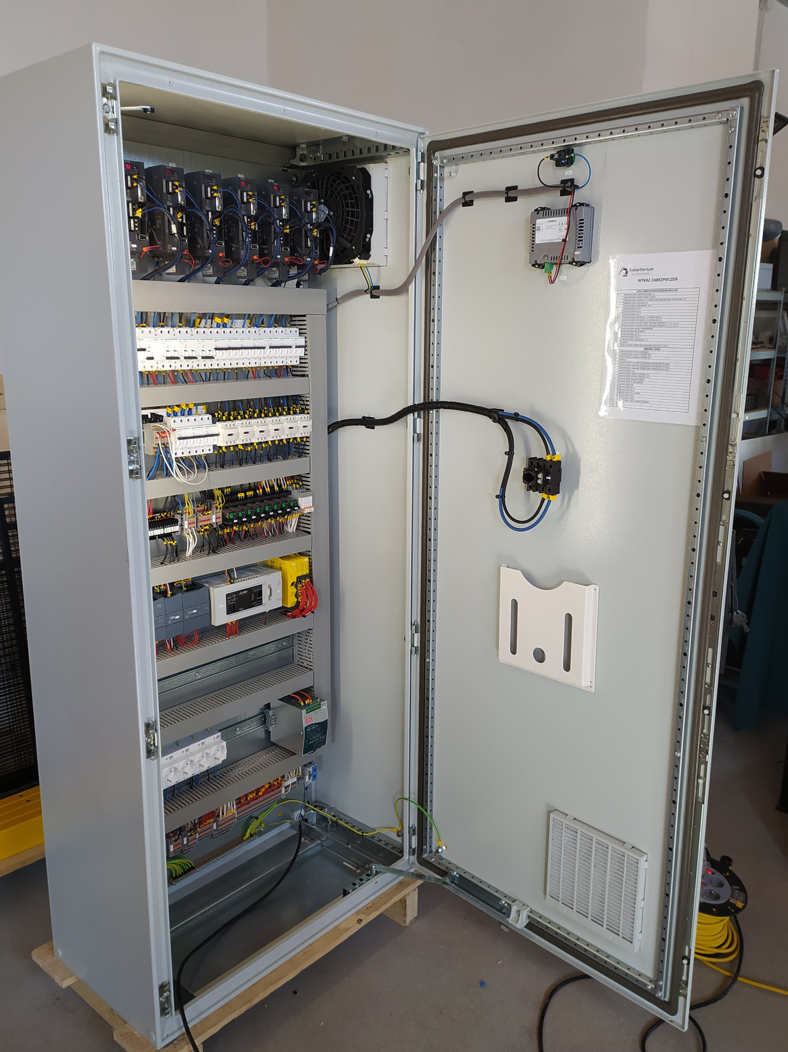 Otwarta szafa sterownicza z widocznymi modułami sterującymi, wentylatorami, okablowaniem i listwą zasilającą, z dokumentacją techniczną na wewnętrznej stronie drzwi.
