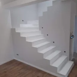 Białe, pomalowane schody w minimalistycznym wnętrzu z brązową podłogą, widoczne otwory na gniazdka elektryczne w ścianie.