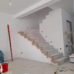 Klatka schodowa w trakcie przygotowania do malowania: schody zabezpieczone taśmą malarską, ściany wstępnie wyszpachlowane, widoczne narzędzia i puszki z farbą.