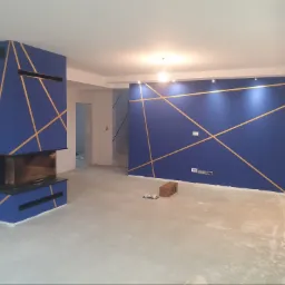 Niebieska ściana z geometrycznym wzorem z taśm w kolorze złota, kominek obudowany w tym samym stylu, surowa podłoga w trakcie prac wykończeniowych, widoczne narzędzia malarskie.