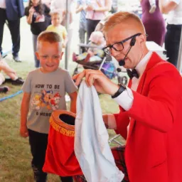 Mężczyzna w czerwonej marynarce i okularach, z mikrofonem, wykonuje sztuczkę magiczną z kapeluszem i chustą, obserwuje go uśmiechnięty chłopiec w koszulce z motywem z bajki Psi Patrol.