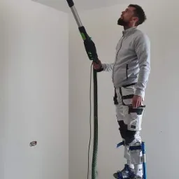 Pracownik na szczudłach szlifuje sufit za pomocą szlifierki z odkurzaczem, widok z dołu.