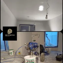 Sprzęt do natryskowego nakładania farby marki Graco Mark V HD z wiadrem farby Brilux Superlux 3000 w pomieszczeniu z pomalowanymi ścianami i sufitem, okno zabezpieczone folią.