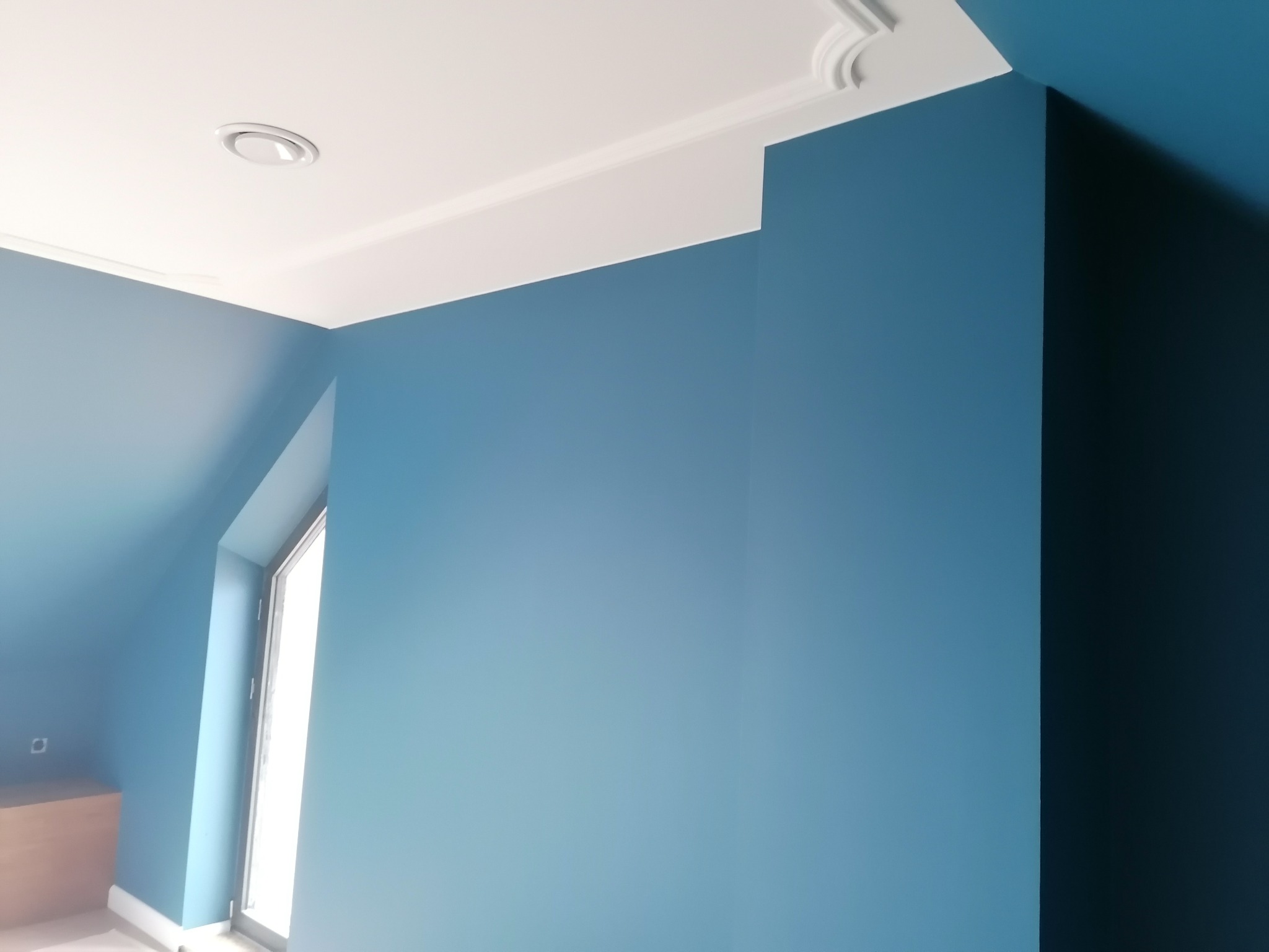 Świeżo pomalowane na niebiesko poddasze z oknem, białą listwą sufitową z dekoracyjnym wykończeniem i okrągłym nawiewnikiem wentylacyjnym.