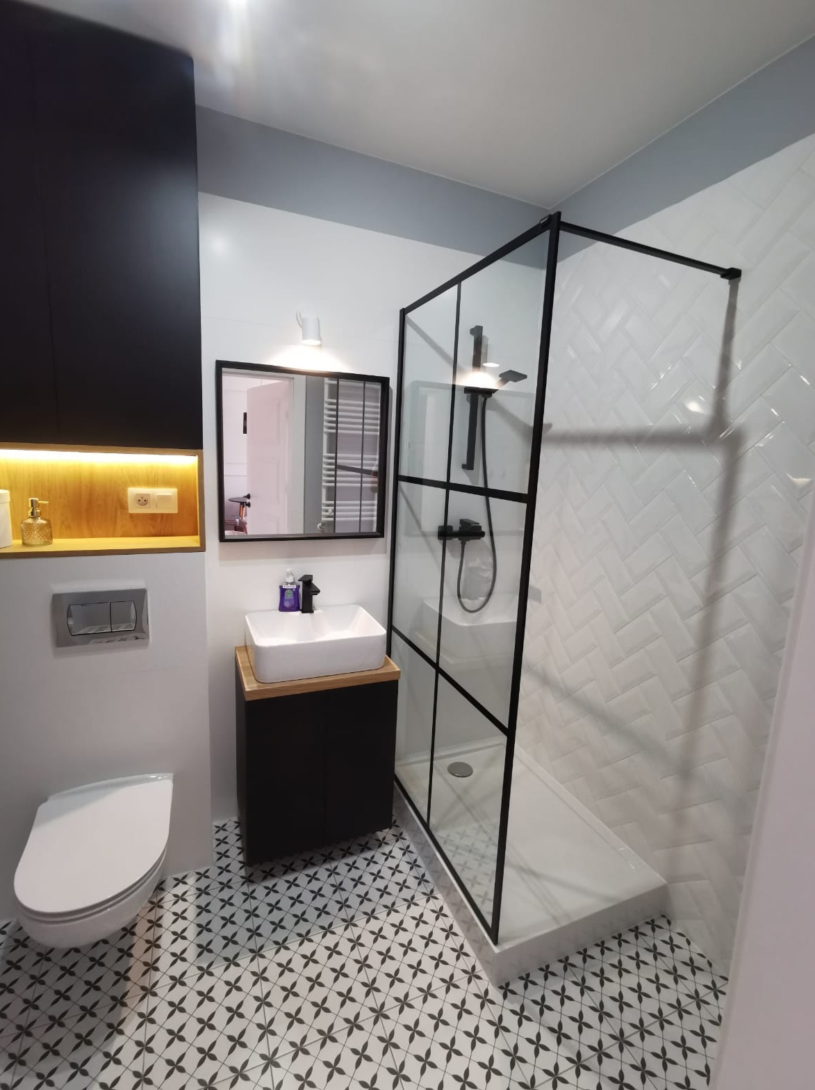 Nowoczesna łazienka z kabiną prysznicową typu walk-in z czarnymi profilami, białą umywalką nablatową na czarnej szafce, lustrem w czarnej ramie oraz podłogą w biało-czarne wzory.
