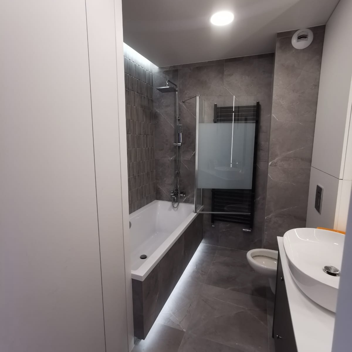 Nowoczesna łazienka z wanną obudowaną szarą płytką z podświetleniem LED, szklanym parawanem nawannowym, grzejnikiem drabinkowym i umywalką nablatową na tle szarych płytek imitujących marmur.